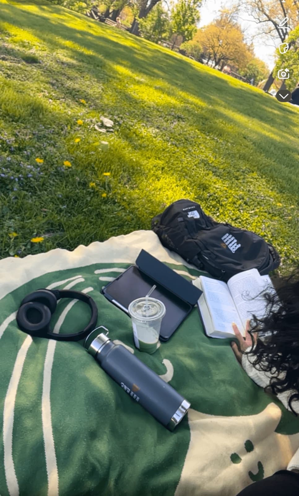 A picnic blanket on the grass with a book, headphones and a drink