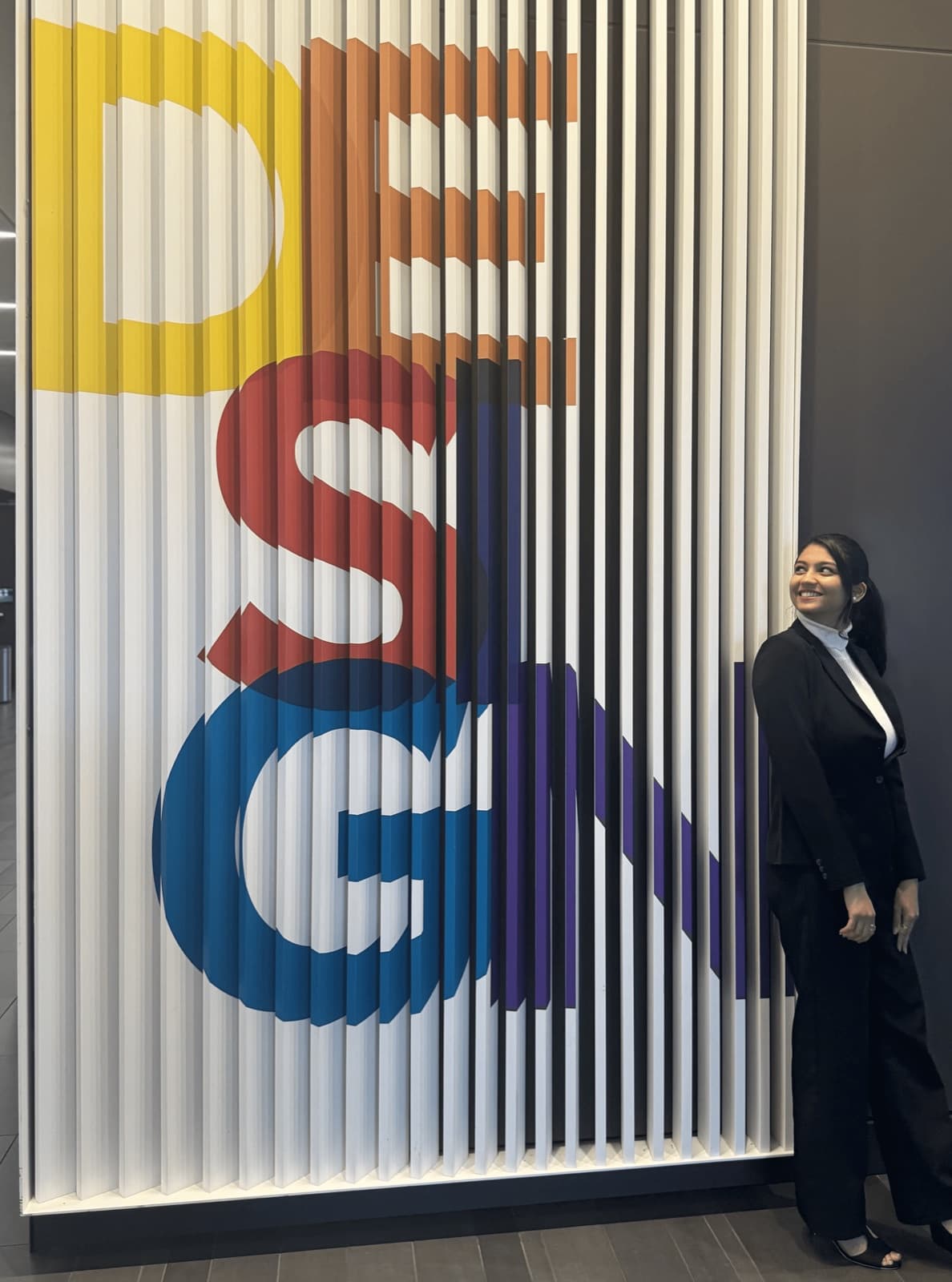 Heena standing beside a large wall sign that reads DESIGN